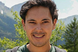 Headshot of a smiling young man standing a forest with mountains in the backgraound