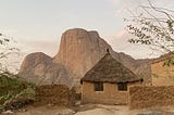 A mud and grass thatched hut sits on a hill in front of unique mountains.