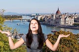 long-haired young person lifts palms to sky and grins. behind her is the Budapest skyline on the water.