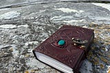 A richly embossed burgundy leather journal with a pentacle design, turquoise gemstone, and beautiful old-style clasp rests on a weathered stone surface on a beach in Norway, evoking a sense of mystery, ritual, and ancient wisdom.