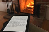 Olive green velvet chair with a kindle on the arm by a lit fireplace. Fall decor ideas for the home in 2024. Photo by Patricia Steffy.