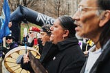 An image of four people standing in a row, taken from profile. Two people are beating drums.