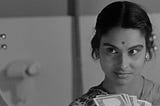 A screen grab from Satyajit Ray film “Charulata”. It is a beautifully styled traditional Indian woman observing herself in a mirror while holding a wad of cash.