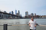 Man wearing a t-shirt and leaning with his back against a railway. Water and the New York skyline is behind him. It is daylight.