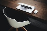 A silver Macbook Pro laptop rests half-closed next to an Apple mouse on a wooden table with a black-and-white chair underneath. Photo by Luca Bravo, via Unsplash.