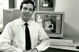 Black and white photograph of a white man with black hair sitting in front of a 1990s computer. he man is wearing a white striped button down shirt and a dark necktie. The man has a slight smile on his face. His hands are clasped in front of his body.