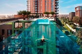 We’re looking at a rooftop pool like no other. It’s made entirely of see-through plastic. It spans the distance between two neighbouring buildings, like a bridge made of water. There’s a woman in the pool, floating on her back. If she were to look down, she’d be able to see the ground, hundreds of feet below her.