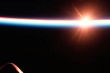 Sunrise viewed from space, with the sun flaring bright orange-white just above Earth’s curved horizon, which glows in a thin band of blue and red against the black of space. Part of a spacecraft’s dark silhouette, including a vertical fin or antenna, is visible in the lower left foreground.