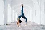 A woman dressed in blue leggings and tank top is doing a backbend amidst a gorgeous series of white archways. She has one leg high in the air.