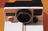 White Polaroid Land Camera with a One Step red button and rainbow running down the middle. This image has an orange background.