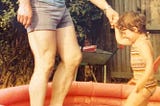 Small child in paddling pool holding her father’s hand
