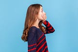 Side view portrait of a woman in a striped casual style sweater covering her mouth with a hand, looking surprised. Indoor studio shot isolated on blue blackground