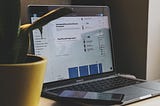 A laptop with the Figma app open and a phone laying on top of the keyboard sits next to a yellow potted plant.