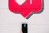 Street art of an Instagram “like” red bubble with heart on a white cement block wall, with a hand holding up a phone beneath it