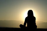 Photograph of a woman sitting cross-legged and meditating outside at sunset.
