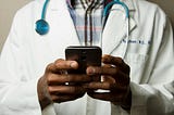 Black Doctor Holding Cell Phone. He has on a plaid shirt under a white coat and has a stethoscope around his neck. His face is out of the frame.