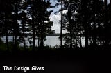 A cloudy sky shines through the darkness of a forest while it is gracefully placed above a lake.