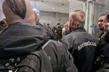 A photo of several men in black jackets, shot from behind. One man with a partly shaved head and a head tattoo wears a jacket with the word “Antisöcial” on the back.