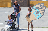 Black mother pushing stroller with toddler, standing next to a child with a fist that says, “Free us 23 million”