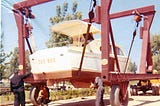 White cabin-cruiser boat that says “Cee Bee” on the back — being hoisted onto a trailer. A man stands at the back of the boat and trailer while another man is working on the right side and another man is squatting down underneath.