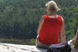 Woman with her back to the camera in a red sleeveless shirt sits on the edge of a cliff looking down at trees.