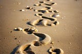 The image depicts a series of footprints on the beach, showcasing the journey of growth and progress. Starting from the left, the footprints begin small, representing a child’s tiny feet, and gradually increase in size as they move towards the right. The smallest footprints have a tentative, faltering quality, while the larger ones become more confident and bold. The footprints lead to a large, adult-sized footprint at the far right, symbolizing the transition from childhood to adulthood. The be