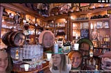 Image shows close up of bar counter, crowded with drinks and cups and bottles and plates, overlayed with bubbles of faces clearly post-edited. Bottom right of image contains text: “ Inspired by The Beanery (1965) by Edward Kienholz”