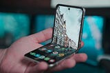 A hand holding a foldable smartphone with the home screen displayed. There is a picture of houses on a street as the background image on the phone.