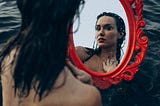 A beautiful woman sitting on the water looking at her reflection at a red mirror.