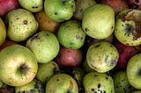A pile of apples, mostly green apples with some red apples, which have worm holes, dark spots, or other blemishes.