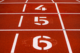 Photo Credit: Photo by Markus Spiske on Unsplash “track and field, red track with white lines” Color photo of a red clay-type of ground for running track and field. I combined two photos by Markus Spiske to create one photo. Top of photo is just the brick red track with white lines. Bottom is a similar colored track with numbers 4–5–6 in white displayed from top to bottom in numerical order.