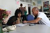 Two people sitting at a table with the older person pointing to an open notebook in front of the younger person. Most likely coaching her on how to do something.