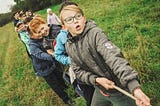 A line of young kids tugging on a rope in a green field.