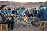A group of happy young people gathered around a campfire. In the background some leafy trees, then further back a rocky line of cliffs with the sun low behind.