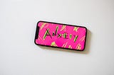 A mobile phone with Pink and background and Anxiety written in black and florescent green strokes.