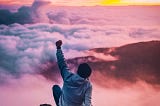 Man sitting on top of a mountain, seeing clouds and raising his fists