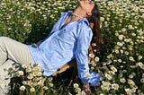 Woman wearing light blue shirt and light grey trousers, lying backwards in a field of daisies.