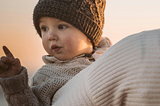 Toddler is bundled in crocheted hat and knit sweater and looking over mom’s shoulder who is carrying him and walking away from the camera. They are both bundled for the snow.