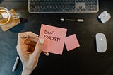 Manicured hand holding a post it reading “Don’t forget” above a computer keyboard and mouse