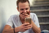 Young man looking at phone and covering mouth as he laughs in excitement.