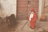 The picture shows a black dog and a 2-year-old baby girl, dressed in a warm red winter outfit, holding a stick