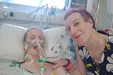 Abbie posing for the camera at Fae’s bedside, hand on Fae’s shoulder. Fae is breathing through medical equipment.