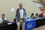 A man in a suit standing and facing the camera in front of a booth outside a convention.