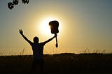 Silhouette of a man holding out his arms, one of which is holding an acoustic guitar against a sunset.