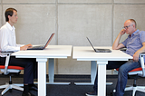 two men sitting across from each other at an office with laptops and one is staring creepily at the other one