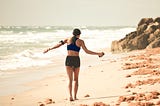 Young Woman walking along a rocky beach, wearing sport shorts and a sports bra, with a phone and headphones. Dark long hair in a low ponytail. Daytime.