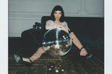 Woman sits on the floor behind a giant disco ball, looking solemn.
