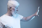 A serious mature bearded male with gray hair doing protective gesture while standing in a white light