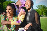 A trio of young black women laugh and embrace while sitting outside.