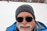 Selfie of the author, John Carney, wearing a stocking cap and heavy winter coat and standing in front of his apartment building. The lawn is covered with snow. Carney’s light-sensitive eyeglass lenses have fully darkened.